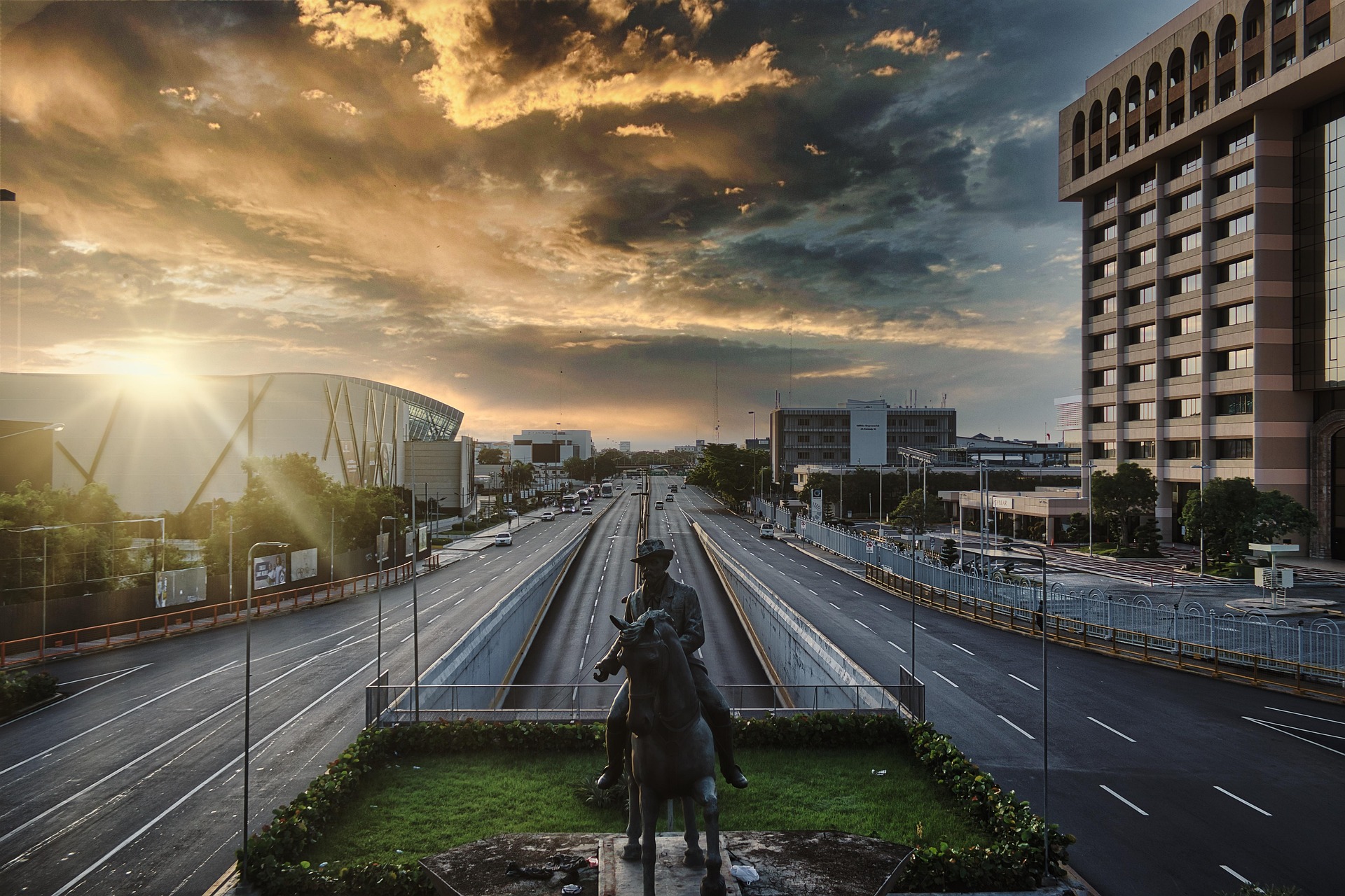 Vista aérea de una gran avenida de múltiples carriles en Santo Domingo al atardecer. En primer plano, una estatua ecuestre de bronce sobre una glorieta con jardín. A los lados se aprecian edificios de varios pisos, vegetación y vehículos. El cielo presenta nubes dramáticas iluminadas por la luz dorada del sol poniente en el horizonte izquierdo.