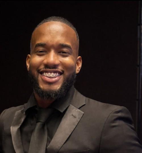 Fotografía de retrato profesional de un hombre joven con barba corta y cuidada, sonriendo a la cámara. Viste un traje formal negro con camisa negra y corbata a juego. El fondo es oscuro y neutro, resaltando una expresión de confianza y amabilidad.