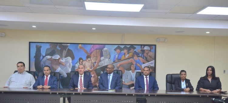 Fotografía panorámica de la mesa de honor en el Salón del Consejo Universitario, donde aparecen siete autoridades académicas sentadas en fila frente a un mural artístico colorido que adorna la pared de fondo. De izquierda a derecha, se encuentran el Director del Instituto de Física vistiendo una chacabana gris; el Decano de la Facultad de Ciencias con traje azul marino y corbata roja; el Ministro del MESCyT con traje oscuro y corbata a rayas rojas; el Rector de la UASD, ubicado en el centro, con traje oscuro y corbata azul celeste; el Vicerrector de Investigación y Postgrado con corbata morada; la Directora de la Escuela de Física con chaqueta oscura y camisa blanca; y la Coordinadora del Programa Doctoral vestida de negro, todos manteniendo una postura formal y mirando hacia el frente.
