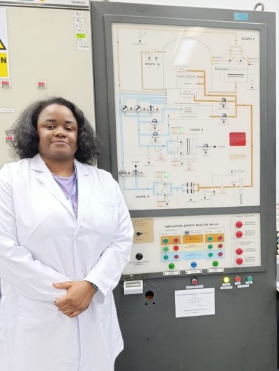 "En la imagen se ve a la investigadora Mariella Mateo Villanueva de pie en un entorno técnico, mirando hacia la cámara con una leve sonrisa y las manos entrelazadas. Lleva puesta una bata blanca de laboratorio sobre una blusa lila y pantalones negros. Detrás de ella se encuentra un gran panel vertical de color gris, identificado con el texto 'VENTILACION EDIFICIO REACTOR RP-10'. Este panel contiene un diagrama esquemático con líneas de flujo, indicadores luminosos y una botonera con controles rojos, verdes y azules. A la derecha de la imagen, sobre un escritorio, se observa un monitor de computadora que muestra gráficos de monitoreo, junto a un cuaderno de notas y un extintor rojo montado en la pared del fondo."